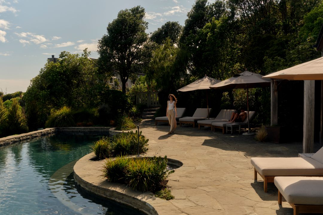 An outdoor pool surrounded by greenery with a wooden lodge, shaded lounge chairs, and a stone patio with lush plants.